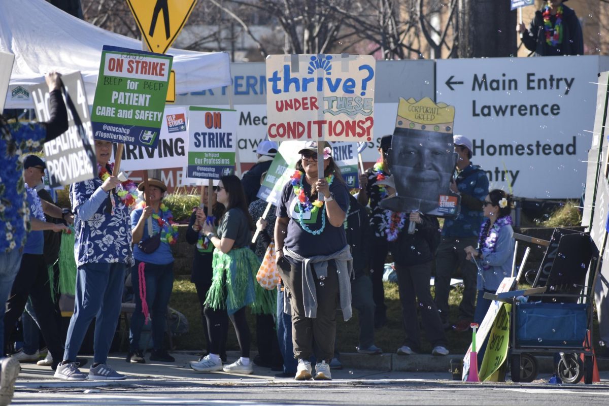 A smiling protester within the crowd holds a sign containing the Kaiser Permanente logo and reading "Thrive? Under these conditions?" on the sidewalk in front of Kaiser Permanente Santa Clara Medical Center on Feb. 2.